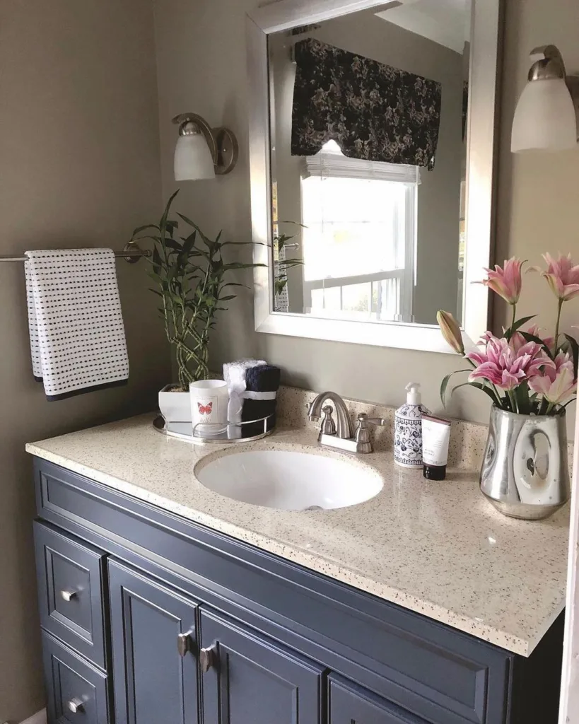 A Grey Bathroom Vanity With Beige Granite Counters And Two Plants As Decor