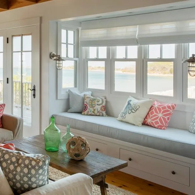 31 Coastal Beach-Themed Living Room Ideas 21 A Beach House Window Nook With Seating And A Sofa Looking Out The Windows To The Ocean