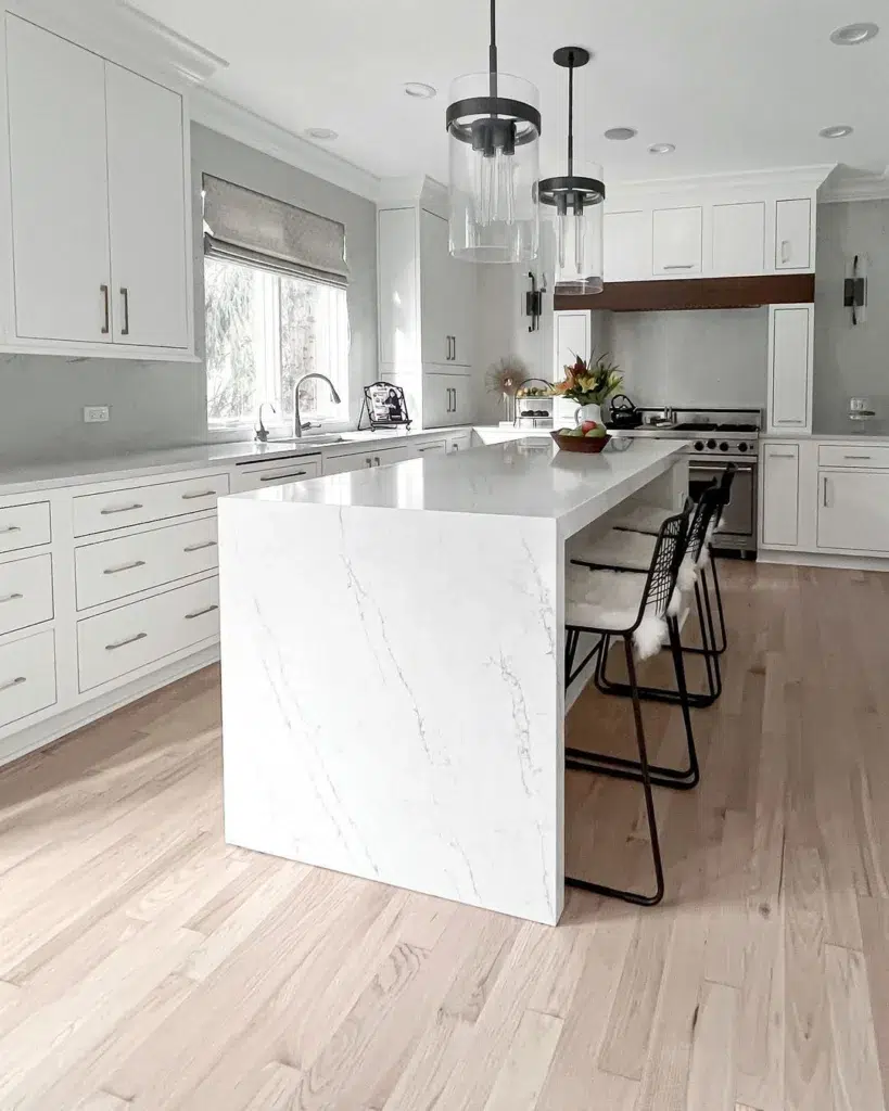 A White Kitchen With Silver Hardware And A Large Waterfall Kitchen Island