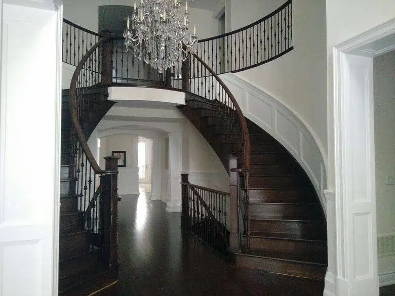 Double Curved Staircases With Dark Brown Treads And Risers And A Chandelier In The Center Of The Room