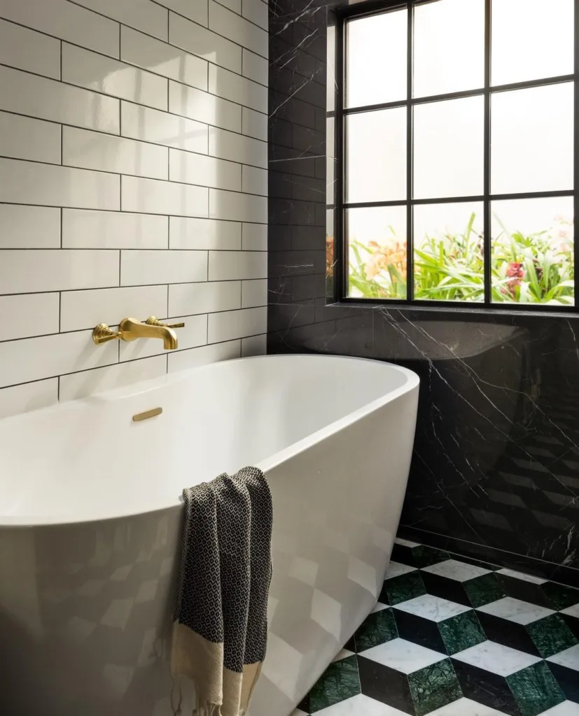 A Sleek And Elegant Bathroom Featuring A Black, White, And Green Marble Hexagonal Tile Pattern On The Floor, Creating A Luxurious And Sophisticated Look. A White Tub Is The Centerpiece Of The Space, Providing A Comfortable And Relaxing Spot For Bathing. White Tiles Are Also Present On The Floor, Adding To The Clean And Minimalist Aesthetic. A Black-Framed Window Brings Natural Light Into The Space, Providing A Contrast To The Cool-Toned Marble Tiles. 