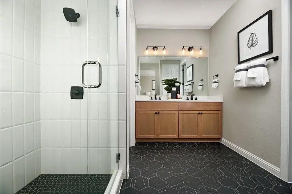 A Chic Bathroom With White Walls And A Striking Black Hexagonal Tile Pattern On The Floor, Creating A Bold And Stylish Contrast. Wooden Cupboards Provide Storage And A Natural Touch, While A Painting Of Leaves On The Wall Adds A Pop Of Color And A Touch Of Artistry To The Room. The Bathing Area Is Enclosed With Glass, Providing A Sleek And Modern Touch. The Hexagonal Tiles Have A Matte Finish, Adding A Subtle Texture To The Space.