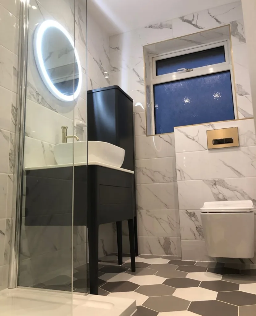 A Luxurious Bathroom With White Marble Walls That Add Elegance And Grandeur To The Space. The Floor Features A Striking Black, Grey, And White Hexagonal Tile Pattern, Creating A Bold And Stylish Contrast. A Lighting Mirror Adds A Modern Touch And Provides Functional Lighting For The Vanity. A Window Lets In Natural Light, Adding Brightness And Warmth To The Room. The Tiles Have A Polished Finish, Reflecting Light And Adding To The High-End Feel Of The Space. 