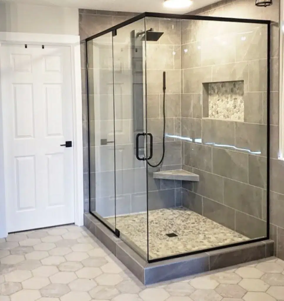 A Minimalist And Modern Bathroom Featuring Cream-Colored Hexagonal Tiles On The Floor, Creating A Clean And Sleek Look. A Glass Partition Separates The Bathing Area, Adding A Sense Of Openness And Modernity To The Space. The Door Is White, Providing A Crisp And Clean Contrast. The Hexagonal Tiles Have A Polished Finish, Reflecting Light And Adding To The Modern And Sophisticated Vibe Of The Room. 