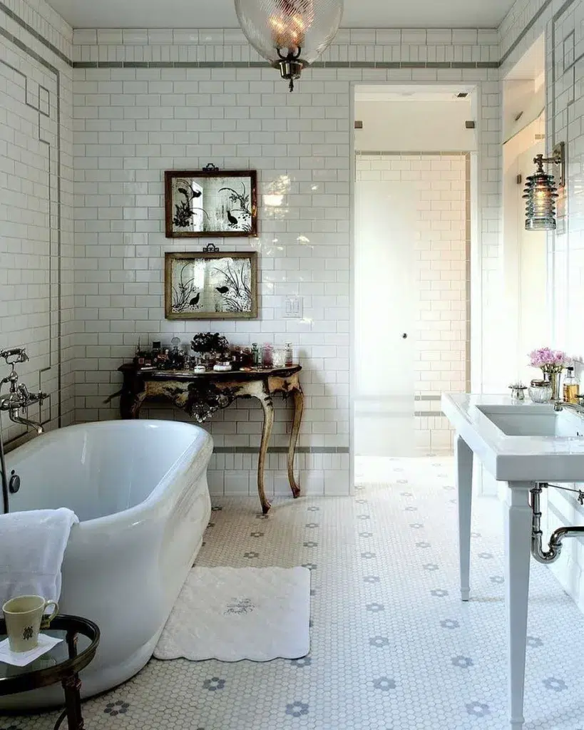 A Charming And Cozy Bathroom Featuring A Small Hexagonal Tile Pattern In Shades Of White And Grey On The Floor, Creating A Subtle And Classic Design. A White Tub Is Situated In The Center Of The Room, Surrounded By The Hexagonal Tiles, Providing A Relaxing And Comfortable Space To Unwind. White Tiles Line The Walls, With A Painting On The Wall Adding A Pop Of Color And Interest To The Space. 