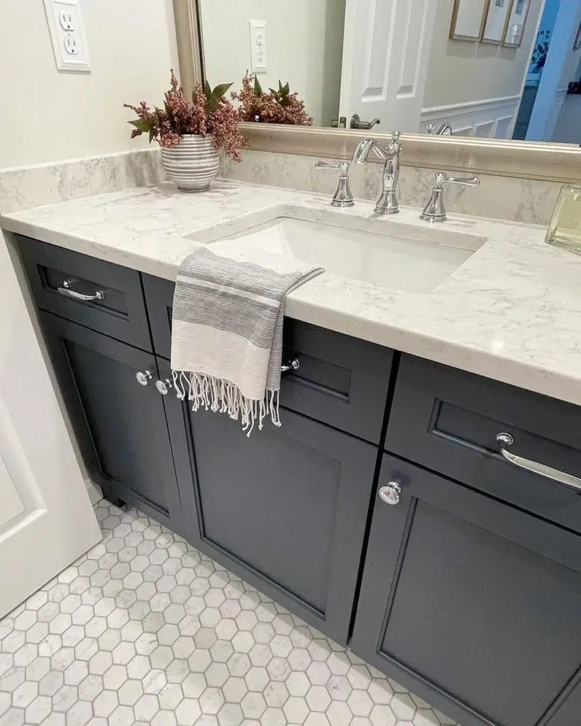 A Modern Bathroom Featuring A White Hexagonal Tile Pattern On The Floor And Black Cupboards, Creating A Sleek And Stylish Contrast. The Cupboards Are Topped With A White Marble Countertop, Adding A Touch Of Luxury And Elegance To The Space. A Vase Of Flowers Adds A Pop Of Color And A Natural Touch. The Rectangular Mirror Adds A Modern And Functional Touch, Reflecting Light And Providing A Practical Space For Grooming.
