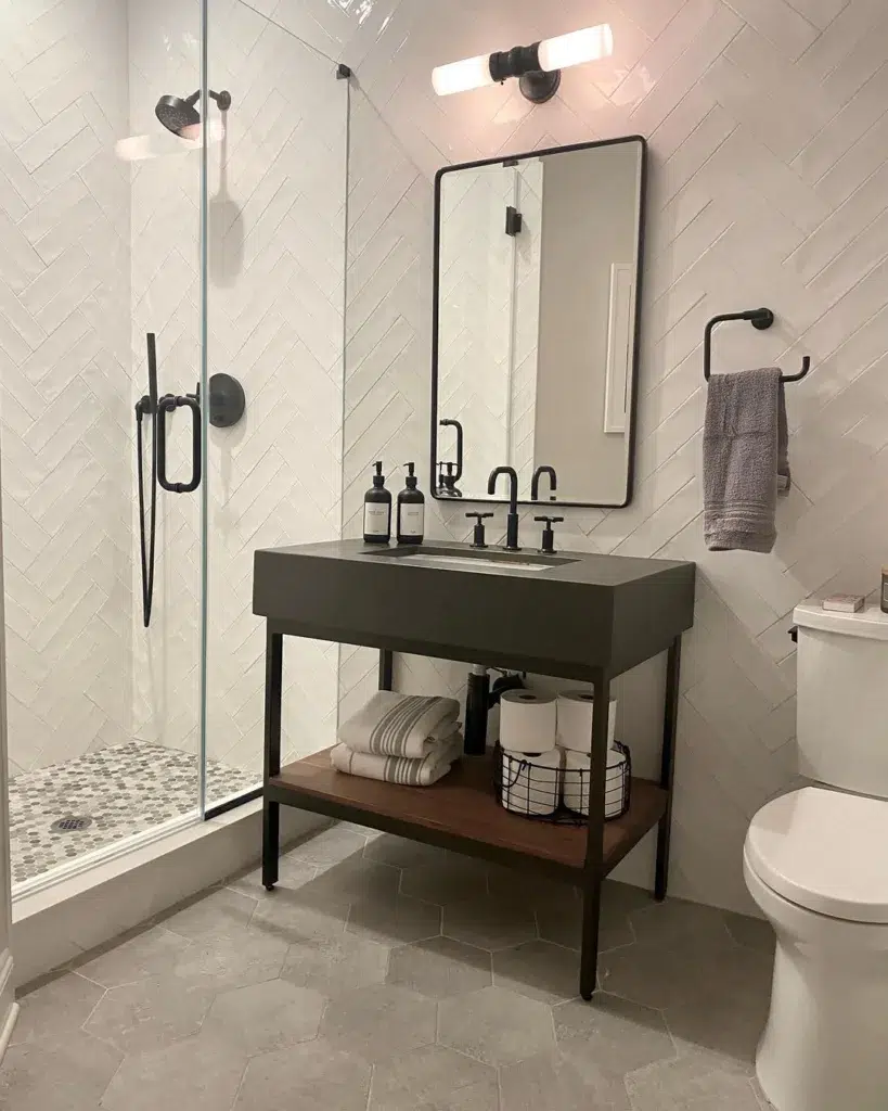 A Modern And Sleek Bathroom Featuring A Grey Hexagonal Tile Pattern On The Floor And White Tiles With A Black Border, Creating A Clean And Contemporary Look. A Mirror On The Wall Is Accompanied By A Table In Front, Providing A Practical Space For Grooming. Shampoo, Conditioner, And A Black Tap Are Within Easy Reach, Adding To The Functionality Of The Space. 