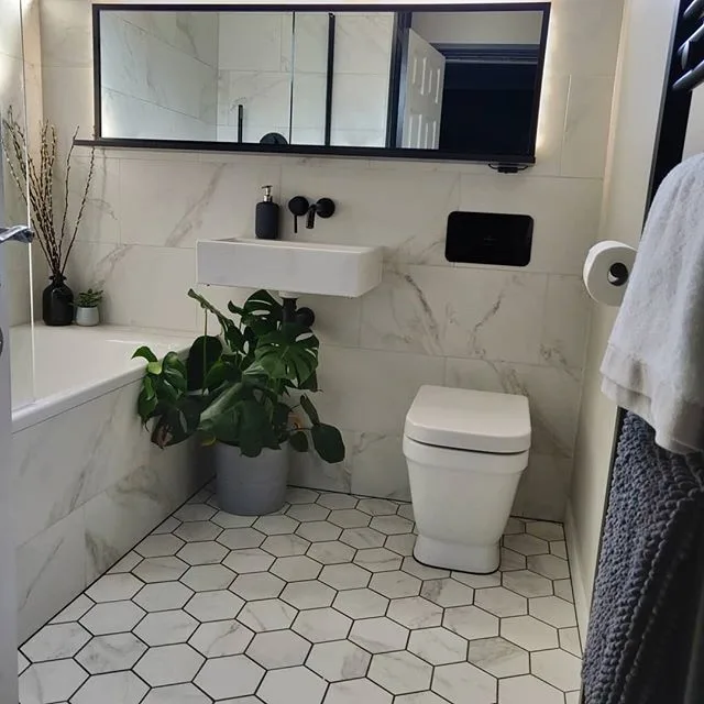A Luxurious Bathroom Featuring A White Hexagonal Tile Pattern On The Floor And White Marble Walls, Creating A Sense Of Opulence And Grandeur. Plants Add A Natural Touch And A Pop Of Greenery To The Space. The Bathtub Is A Focal Point, With A Sleek And Modern Design That Adds To The Luxurious Feel Of The Room. A Black Mirror Provides A Striking Contrast, Adding A Touch Of Drama To The Space. The Hexagonal Tiles Have A Polished Finish, Reflecting Light And Adding To The High-End Feel Of The Room.