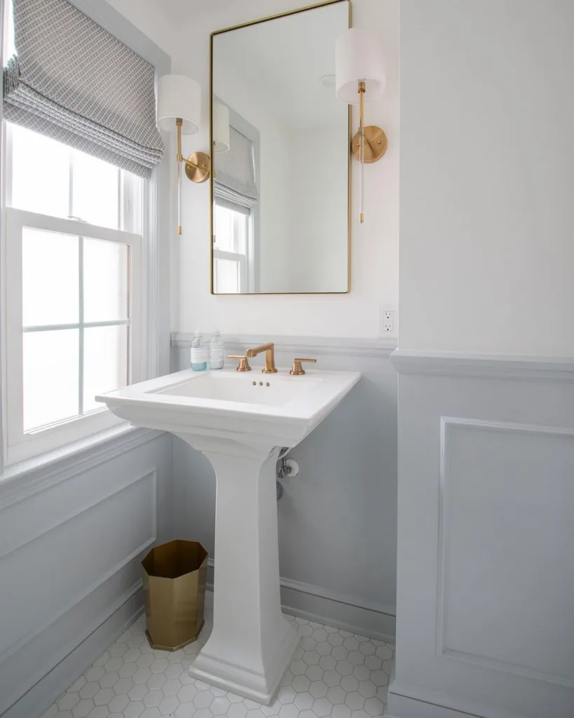 A Bright And Airy All-White Bathroom Featuring White Tiles On The Walls And A White Sink, Creating A Clean And Refreshing Look. The Hexagonal Tile Pattern On The Floor Is Also White, Adding To The Sense Of Spaciousness And Lightness In The Room. A Bathroom Mirror With A Border Of Golden Trim Is Located Above The Sink, Adding A Touch Of Elegance And Refinement To The Space. 