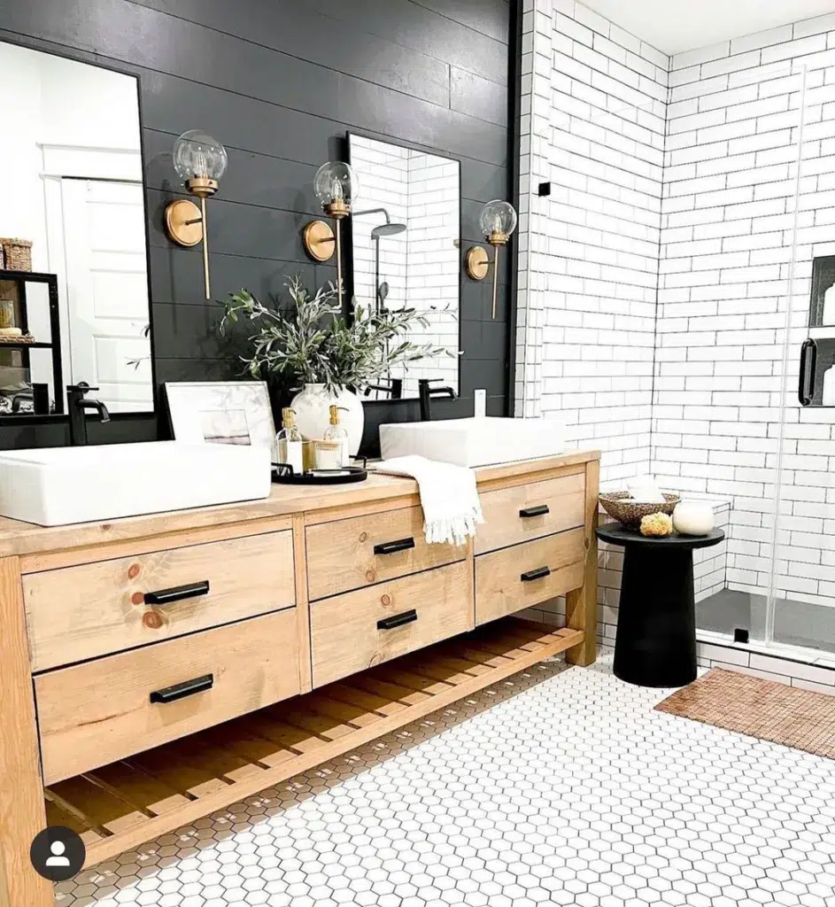 A Modern And Minimalist Bathroom Featuring White Tiles On The Walls And A Black-Framed Wall Mirror, Creating A Sleek And Stylish Look. Three Lanterns Hang Above The Space, Providing Warm And Inviting Lighting. The Hexagonal Tile Pattern On The Floor Is A Clean And Crisp White, Adding To The Minimalist Feel Of The Room. Wooden Cupboards Are Located On The Right Side Of The Space, Providing Storage And Adding A Touch Of Warmth And Texture