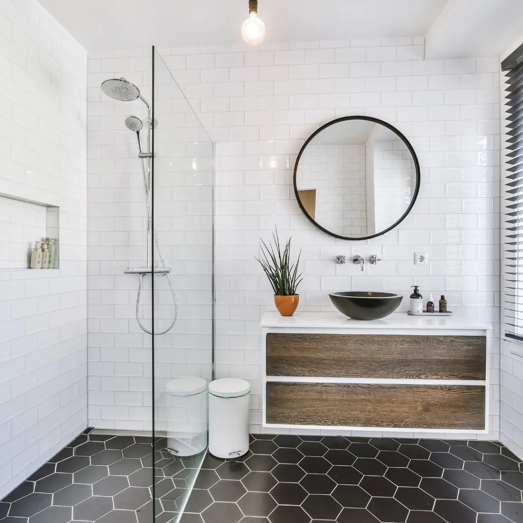 A Modern Bathroom With White Tiles On The Walls And A Striking Black Hexagonal Tile Pattern On The Floor, Creating A Bold And Stylish Contrast. The Bathing Area Is Enclosed With Glass, Providing A Sleek And Modern Touch. A Window On The Left Side Of The Room Lets In Natural Light, Adding Brightness And Warmth To The Space. The Wooden Cupboards Are Decorated With Soap, Shampoo, And Conditioner, Adding A Natural And Organic Touch To The Room. A Small Potted Plant Adds A Touch Of Greenery, Bringing Life And Color To The Space