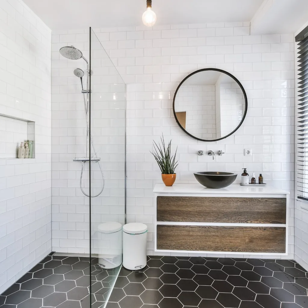 38 Bathrooms With Hexagon Tile Floors 10 A Modern Bathroom With White Tiles On The Walls And A Striking Black Hexagonal Tile Pattern On The Floor, Creating A Bold And Stylish Contrast. The Bathing Area Is Enclosed With Glass, Providing A Sleek And Modern Touch. A Window On The Left Side Of The Room Lets In Natural Light, Adding Brightness And Warmth To The Space. The Wooden Cupboards Are Decorated With Soap, Shampoo, And Conditioner, Adding A Natural And Organic Touch To The Room. A Small Potted Plant Adds A Touch Of Greenery, Bringing Life And Color To The Space